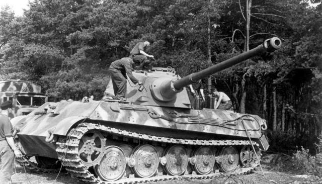 Sort-hvidt foto af en Tiger II med lang kanon og soldater, der står på skroget, i skovkanten